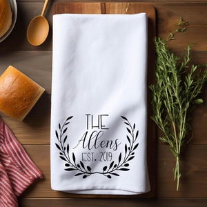 May include: White kitchen towel with a black floral wreath design. The text "THE Allens EST. 2019" is centered within the wreath. The towel is folded and placed on a wooden cutting board, with bread and herbs in the background.
