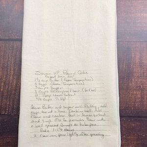 May include: A cream-coloured rectangular kitchen towel featuring a handwritten recipe for Seven-Up Pound Cake in black ink. The text includes ingredients like butter, eggs, sugar, flour, lemon extract, and 7-Up, along with baking instructions.