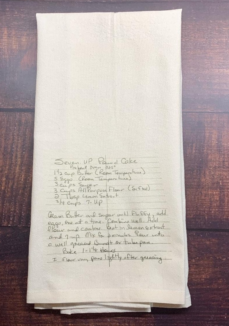 May include: A cream-coloured rectangular kitchen towel featuring a handwritten recipe for Seven-Up Pound Cake in black ink. The text includes ingredients like butter, eggs, sugar, flour, lemon extract, and 7-Up, along with baking instructions.