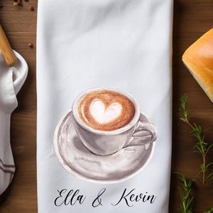 May include: A white kitchen towel with a watercolor illustration of a cup of coffee with a heart design in the foam. The towel has the text 'Ella & Kevin' printed on it.