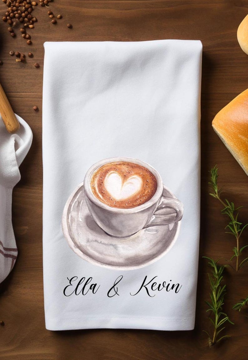 May include: A white kitchen towel with a watercolor illustration of a cup of coffee with a heart design in the foam. The towel has the text 'Ella & Kevin' printed on it.