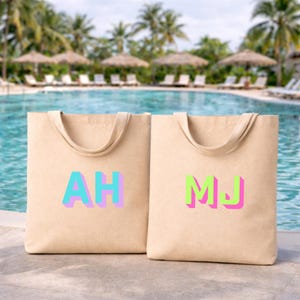 Pode incluir: Duas sacolas bege com iniciais coloridas. Uma sacola tem "AH" em azul, turquesa e rosa, e a outra "MJ" em verde e rosa. As sacolas têm alças combinando e estão em um fundo de piscina.