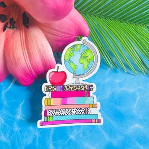 May include: A sticker featuring a stack of six colorful books with various patterns including leopard print and polka dots. A bright red apple sits on top of the books next to a globe with a silver stand. The design has a white border outline.