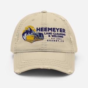 May include: A beige distressed baseball cap with an embroidered logo featuring a yellow bulldozer in front of mountains. The logo reads "Heemeyer Land Clearing & Welding - Est 2004 - Granby, CO".