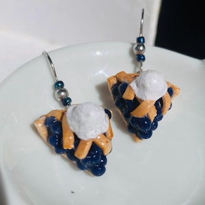 May include: A pair of dangle earrings featuring a blueberry pie slice with a dollop of whipped cream. The earrings are made of polymer clay and have silver hooks.