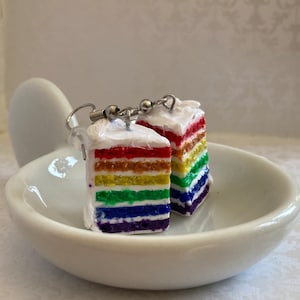 May include: A pair of rainbow layer cake earrings. Each earring is a slice of cake with white frosting and rainbow colored layers. The earrings are made of polymer clay and have silver hooks.