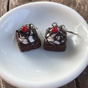 May include: A pair of silver dangle earrings featuring miniature chocolate cake with white frosting and a red cherry on top.