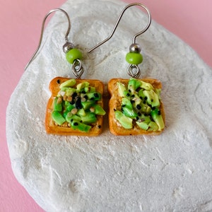 May include: A pair of silver dangle earrings featuring miniature avocado toast. The toast is made of brown clay and topped with green avocado slices and black sesame seeds.
