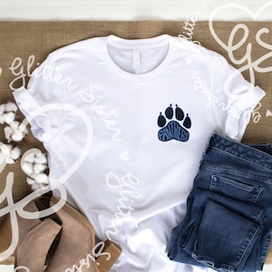 Pode incluir: Camiseta branca com uma pegada azul e a palavra "Panthers" dentro da pegada. A camiseta está sobre uma superfície de juta marrom com um par de botas de tornozelo marrons e calças jeans azuis.