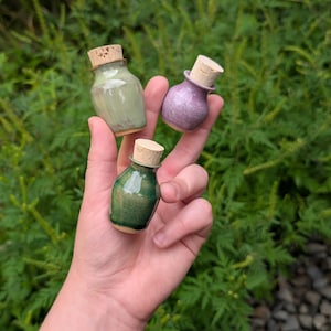 Op de afbeelding: Drie miniatuur keramische flesjes met kurken stoppen. Eén flesje is licht saliegroen, één is diep bosgroen en één is zacht lavendelpaars. Elk flesje heeft een gladde, geglazuurde afwerking en een afgeronde, bolvormige vorm.