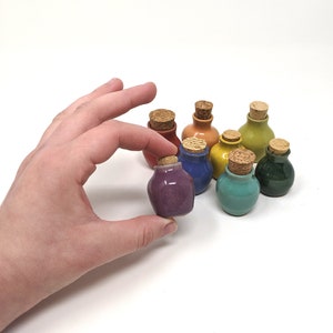 Puede incluir: Una mano sostiene una pequeña botella de cerámica morada con un tapón de corcho. Varias otras botellas coloridas en tonos de rojo, azul, naranja, amarillo, verde y turquesa están dispuestas en el fondo. Las botellas son pequeñas y redondas.