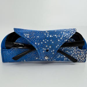 May include: A blue leather eyeglass case with a white speckled pattern, holding a pair of black sunglasses. The case is closed with a silver snap. The sunglasses have black frames.