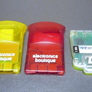Peut inclure: Trois cartes mémoire rétro. Deux sont rectangulaires, translucides et portent l'inscription "electronics boutique" en blanc. L'une est jaune, l'autre rouge. La troisième est transparente avec un circuit imprimé vert et les étiquettes "1 Mb" et "Gamestar".