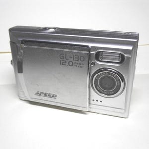 May include: Silver digital camera with the text "GL-130 12.0 Mega Pixel" and "SPEED" printed on the front. The camera lens has a black center and silver ring. The camera has a built-in flash.
