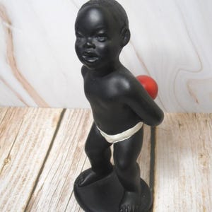 May include: A black figurine of a child holding a red ball behind their back. The child is wearing a white diaper. The figurine is standing on a black oval base.