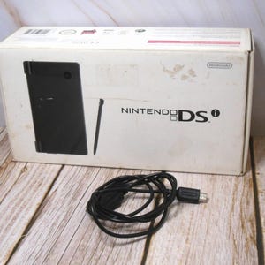 Könnte beinhalten: Eine weiße Nintendo DS-Box mit dem Aufdruck "NINTENDO DS" auf der Vorderseite. Die Box enthält eine schwarze Handheld-Konsole und einen Stift. Ein schwarzes Kabel mit USB-Anschluss liegt vor der Box.
