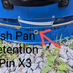 Puede incluir: Un componente met&aacute;lico azul con un orificio circular de color lat&oacute;n para el pasador de retenci&oacute;n de la bandeja de cenizas. La superficie incluye un elemento gr&aacute;fico blanco y el texto "Ash Pan Retention Pin X3" con una flecha roja.