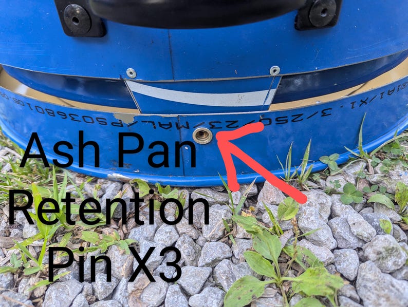 Puede incluir: Un componente met&aacute;lico azul con un orificio circular de color lat&oacute;n para el pasador de retenci&oacute;n de la bandeja de cenizas. La superficie incluye un elemento gr&aacute;fico blanco y el texto "Ash Pan Retention Pin X3" con una flecha roja.