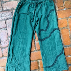 Könnte beinhalten: Waldgrüne Hose mit weitem Bein, elastischem Bund und Kordelzug. Auf dem linken Hosenbein befindet sich eine lange, geschwungene schwarze Schlangengrafik mit texturiertem Schuppenmuster, die vom Oberschenkel bis zum Saum verläuft.
