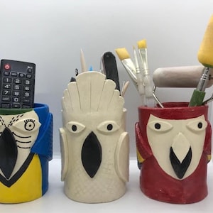 Puede incluir: Tres porta lápices de cerámica con forma de pájaros de dibujos animados. El pájaro azul tiene un pico amarillo y detalles amarillos. El pájaro blanco tiene un pico negro. El pájaro rojo tiene un pico amarillo y detalles amarillos.