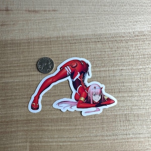 Peut inclure: Un autocollant découpé représentant un personnage d'anime rouge dans une pose dynamique. Le personnage porte un justaucorps rouge avec des détails blancs et a des cheveux roses. Une pièce est à côté de l'autocollant pour la comparaison de taille.