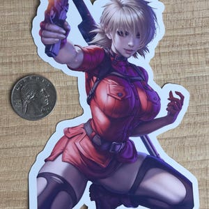 May include: A sticker featuring a woman with blonde hair, wearing a red jumpsuit and holding a gun. She is kneeling with one leg bent and the other extended. The sticker is on a wooden surface.