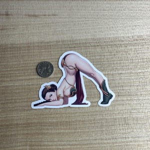 Puede incluir: Una pegatina con una ilustración estilizada de una mujer con un atuendo dorado y verde, con una cortina roja oscura. Está en una pose de yoga, usando botas verdes. Una moneda de veinticinco centavos estadounidense está al lado de la pegatina para comparar el tamaño.