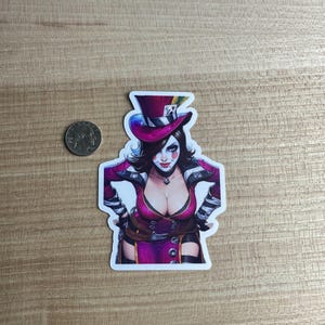 May include: A sticker featuring a stylized illustration of a woman in a purple outfit and top hat. The woman has white face paint, dark hair, and a pendant necklace. A U.S. quarter coin is next to the sticker.