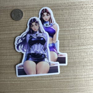 May include: Two die-cut stickers featuring a female character in a superhero costume. The character has long dark hair and is wearing a purple and black outfit. A quarter is next to the stickers for scale.