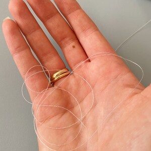 May include: A close-up of a hand holding a length of clear thread. The thread is wrapped around the fingers and is tied in a knot. A gold ring is worn on the middle finger.
