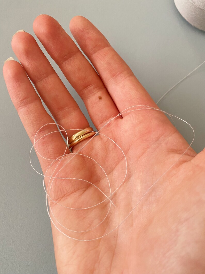 May include: A close-up of a hand holding a length of clear thread. The thread is wrapped around the fingers and is tied in a knot. A gold ring is worn on the middle finger.