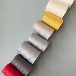 May include: A set of eight spools of thread in various colors, including red, gray, silver, white, yellow, and cream. The spools are arranged in a diagonal line, with the red spool at the bottom and the cream spool at the top.