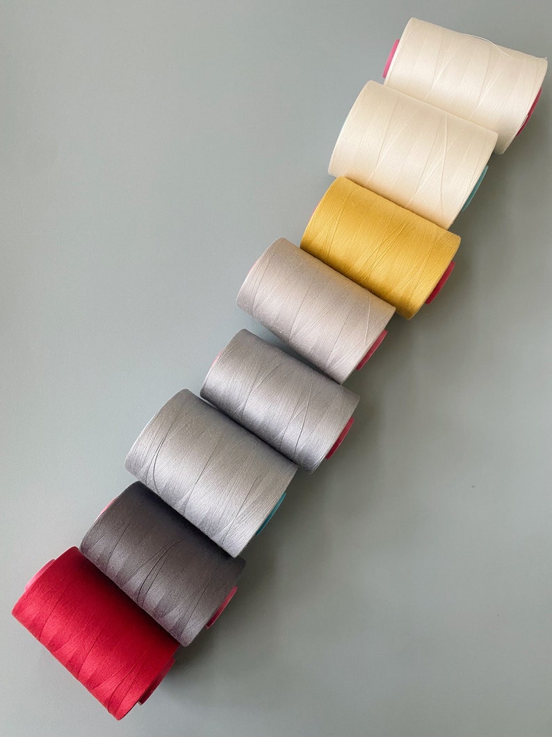 May include: A set of eight spools of thread in various colors, including red, gray, silver, white, yellow, and cream. The spools are arranged in a diagonal line, with the red spool at the bottom and the cream spool at the top.