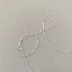 May include: A close-up of a white thread on a light gray surface. The thread is looped and forms a figure eight shape.