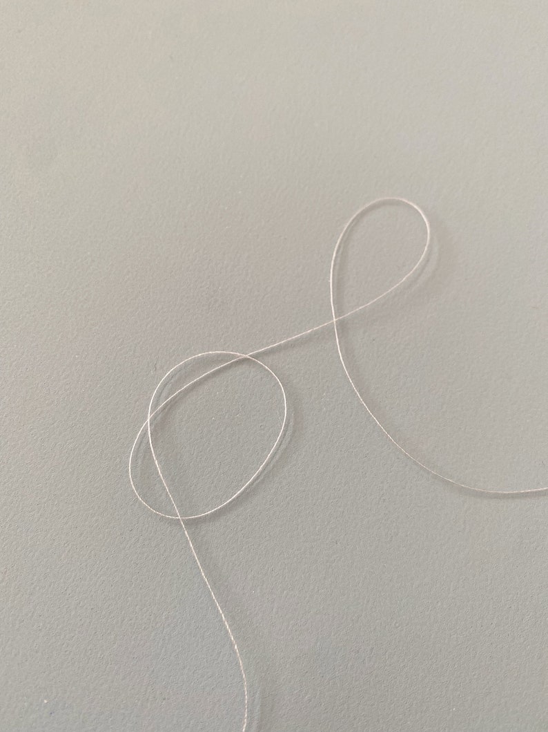 May include: A close-up of a white thread on a light gray surface. The thread is looped and forms a figure eight shape.
