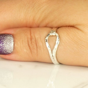 May include: A silver ring with a unique, twisted, open design. The ring is worn on a finger with purple and silver glitter nail polish.