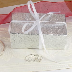 May include: A silver gift box with a white ribbon and bow, sitting on a white surface with a silver bangle bracelet and a pair of silver earrings.