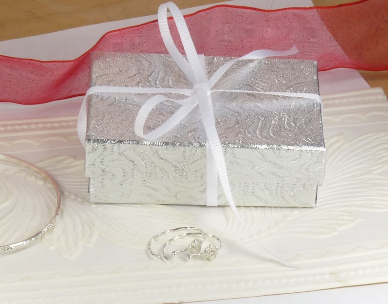 May include: A silver gift box with a white ribbon and bow, sitting on a white surface with a silver bangle bracelet and a pair of silver earrings.