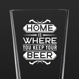 May include: A clear glass pint glass with a white graphic that reads "Home is where you keep your beer".