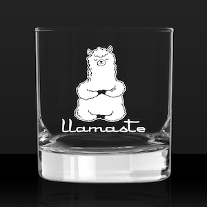 Op de afbeelding: Een helder glazen tumbler met een witte omtrek van een mediterende lama met de tekst "Llama-ste" gedrukt op de bodem.