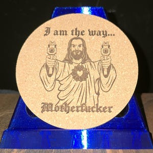 K&ouml;nnte beinhalten: Ein runder beigefarbener Korkuntersetzer mit einer dunkelbraunen Lasergravur einer b&auml;rtigen Figur in Roben, die zwei Handfeuerwaffen h&auml;lt. Der Text "I am the way..." steht &uuml;ber der Figur und "Motherfucker" ist darunter in Schwarz gedruckt.