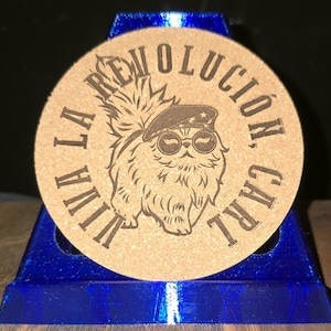 K&ouml;nnte beinhalten: Ein runder beigefarbener Korkuntersetzer mit einer dunkelbraunen, lasergravierten Illustration einer flauschigen Katze, die ein Barett und eine runde Sonnenbrille tr&auml;gt. Der Text VIVA LA REVOLUCION, CARL ist kreisf&ouml;rmig um das Motiv gedruckt.