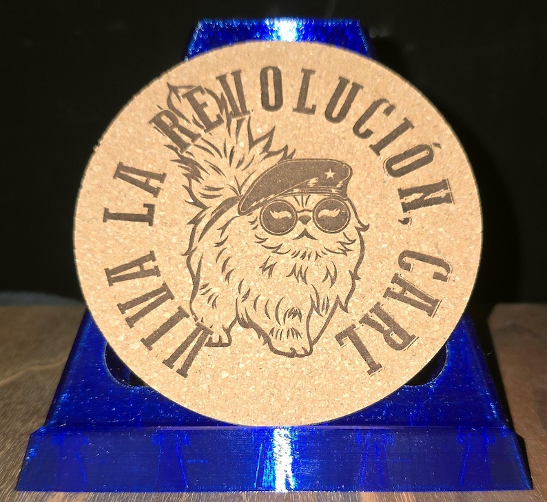 K&ouml;nnte beinhalten: Ein runder beigefarbener Korkuntersetzer mit einer dunkelbraunen, lasergravierten Illustration einer flauschigen Katze, die ein Barett und eine runde Sonnenbrille tr&auml;gt. Der Text VIVA LA REVOLUCION, CARL ist kreisf&ouml;rmig um das Motiv gedruckt.
