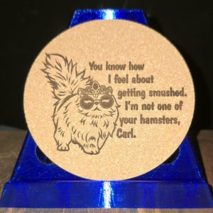 K&ouml;nnte beinhalten: Ein runder, hellbrauner Korkuntersetzer mit einer dunkelbraunen Lasergravur eines flauschigen Katzenmotivs, das eine Krone und eine Sonnenbrille tr&auml;gt. Der Text lautet: You know how I feel about getting smushed. I'm not one of your hamsters, Carl.