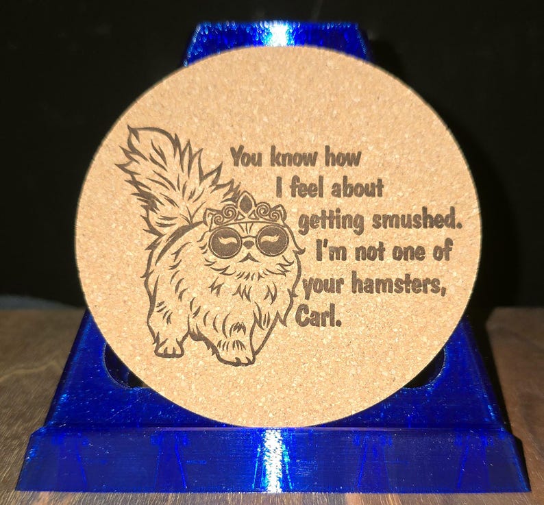 K&ouml;nnte beinhalten: Ein runder, hellbrauner Korkuntersetzer mit einer dunkelbraunen Lasergravur eines flauschigen Katzenmotivs, das eine Krone und eine Sonnenbrille tr&auml;gt. Der Text lautet: You know how I feel about getting smushed. I'm not one of your hamsters, Carl.