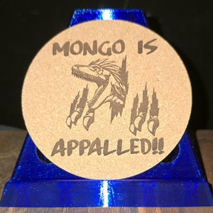 K&ouml;nnte beinhalten: Ein runder, beigefarbener Korkuntersetzer mit einer dunkelbraunen Lasergravur, die einen Dinosaurierkopf zeigt, der aus einem Riss hervorbricht, flankiert von Krallenspuren. Der Text "MONGO IS" steht dar&uuml;ber und "APPALLED!!" darunter.
