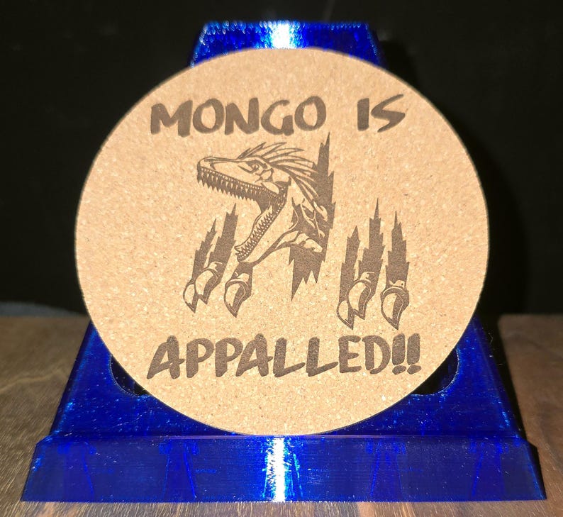 K&ouml;nnte beinhalten: Ein runder, beigefarbener Korkuntersetzer mit einer dunkelbraunen Lasergravur, die einen Dinosaurierkopf zeigt, der aus einem Riss hervorbricht, flankiert von Krallenspuren. Der Text "MONGO IS" steht dar&uuml;ber und "APPALLED!!" darunter.