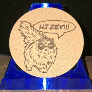 K&ouml;nnte beinhalten: Ein runder, beigefarbener Korkuntersetzer mit einer dunkelbraunen, lasergravierten Illustration einer flauschigen Katze, die eine Krone und eine runde Brille tr&auml;gt. Eine Sprechblase &uuml;ber der Katze enth&auml;lt den Text HI ZEV!!!. Der Untersetzer steht auf einem blauen St&auml;nder.