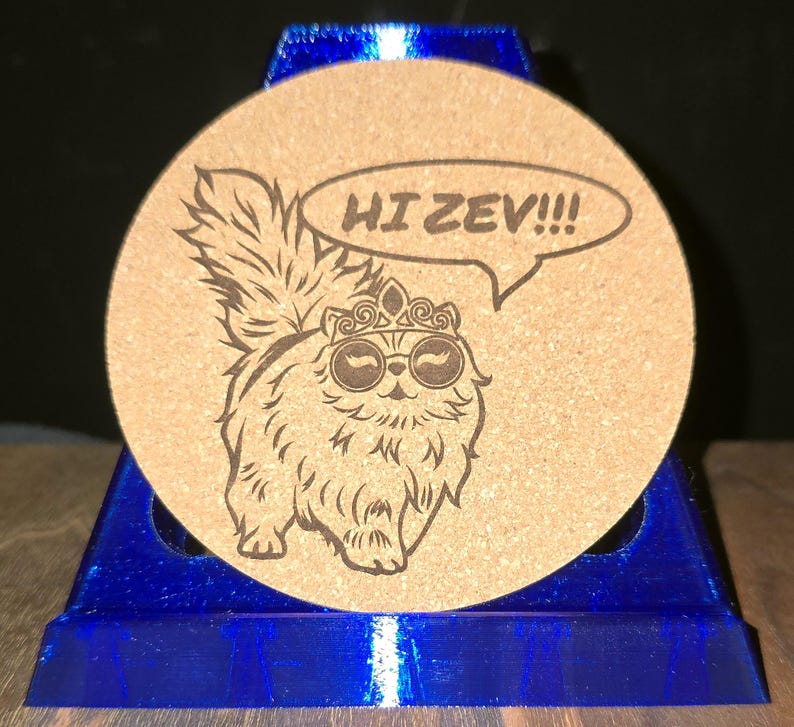 K&ouml;nnte beinhalten: Ein runder, beigefarbener Korkuntersetzer mit einer dunkelbraunen, lasergravierten Illustration einer flauschigen Katze, die eine Krone und eine runde Brille tr&auml;gt. Eine Sprechblase &uuml;ber der Katze enth&auml;lt den Text HI ZEV!!!. Der Untersetzer steht auf einem blauen St&auml;nder.