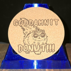 K&ouml;nnte beinhalten: Ein runder, beigefarbener Korkuntersetzer mit einer dunkelbraunen Lasergravur eines flauschigen Katzenmotivs, das eine Krone und eine runde Sonnenbrille tr&auml;gt. Der Text "GODDAMNIT DONUT!!!" ist in fetter Schrift &uuml;ber und unter der Katze eingraviert.